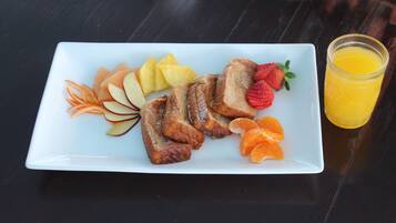 Um prato retangular branco com torradas francesas, fatias de maçã, gomos de laranja e morangos, acompanhado por um copo de suco de laranja sobre uma mesa de madeira escura.