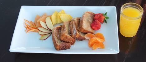 A white rectangular plate with French toast, apple slices, orange segments, and strawberries, accompanied by a glass of orange juice on a dark wooden table.