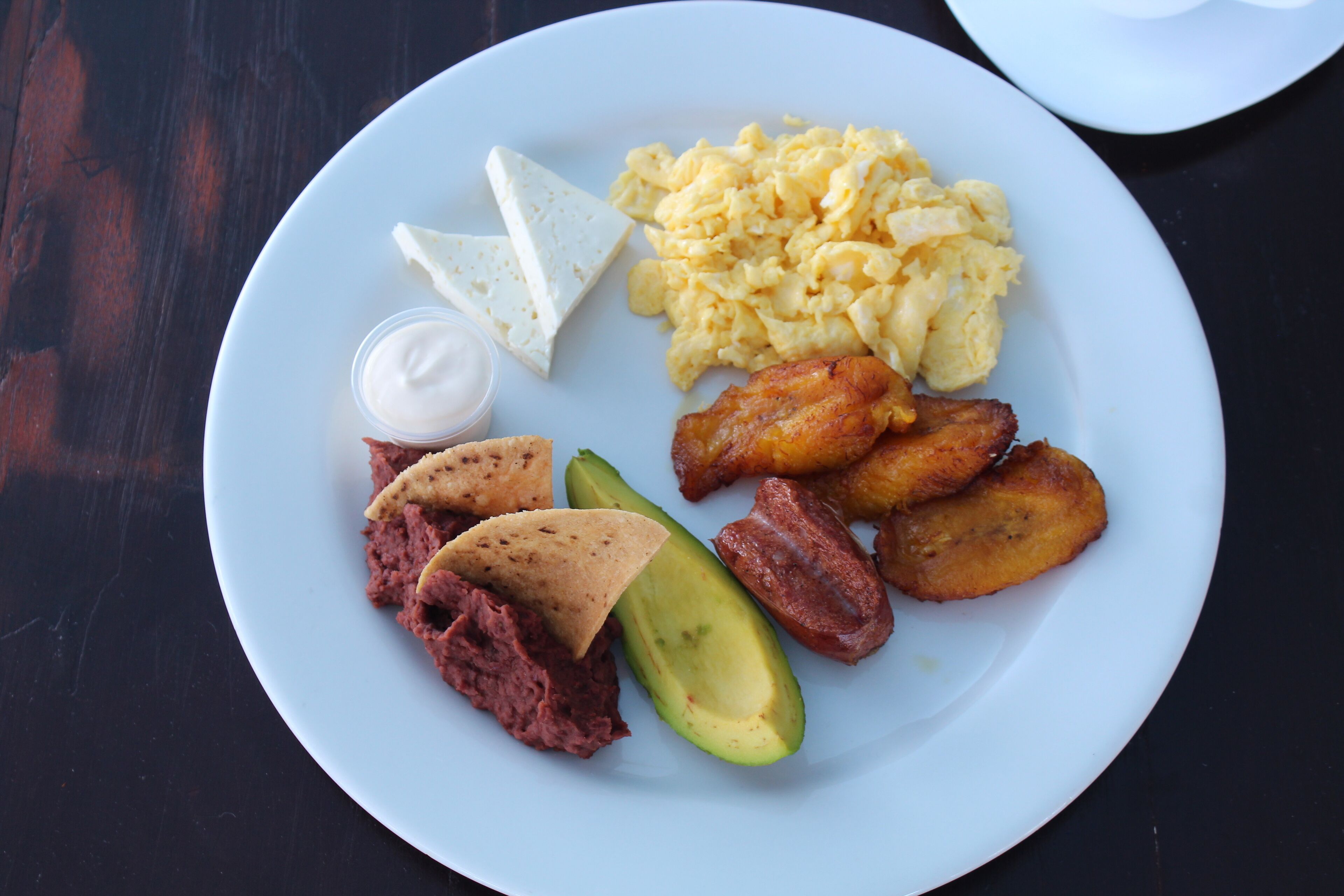 Un plato con huevos revueltos, rebanadas de aguacate, frijoles refritos y plátanos fritos.