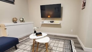 A small, modern room with a flat-screen TV mounted on the wall, a small white table with a lamp, a blue sofa, and a patterned rug.