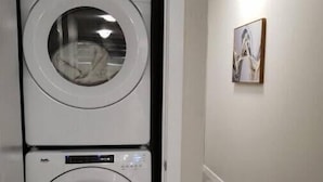 A double wall-mounted washer and dryer unit in a hallway with a light-colored floor and a framed picture on the wall.