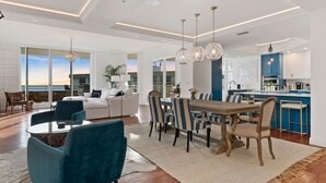 A modern living room with a dining area, large windows, and a kitchen with blue cabinets.