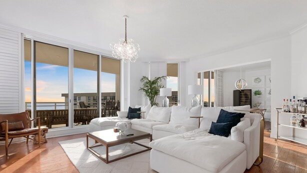 A spacious living room with a white sofa set, a wooden coffee table, and a large window offering a view of the outdoors.