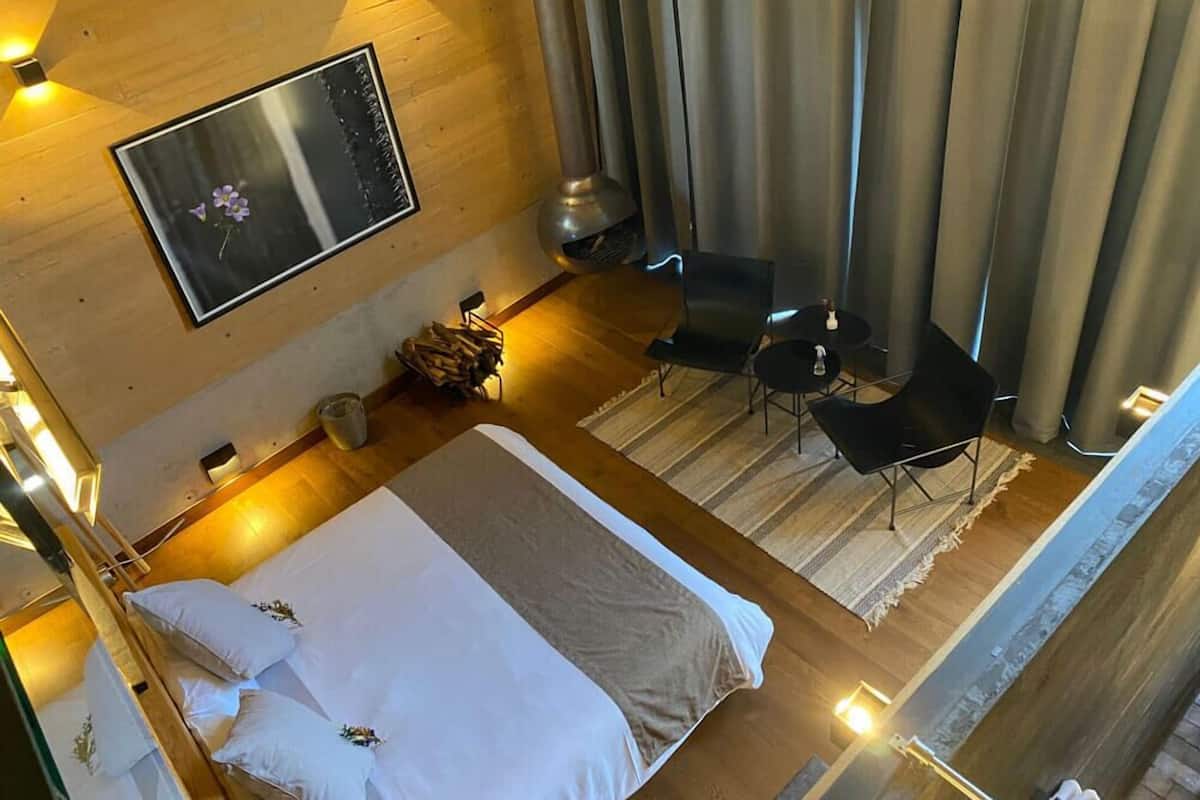 A modern hotel room with a large bed, a small seating area with two chairs, a fireplace, and a large mirror on the wall.