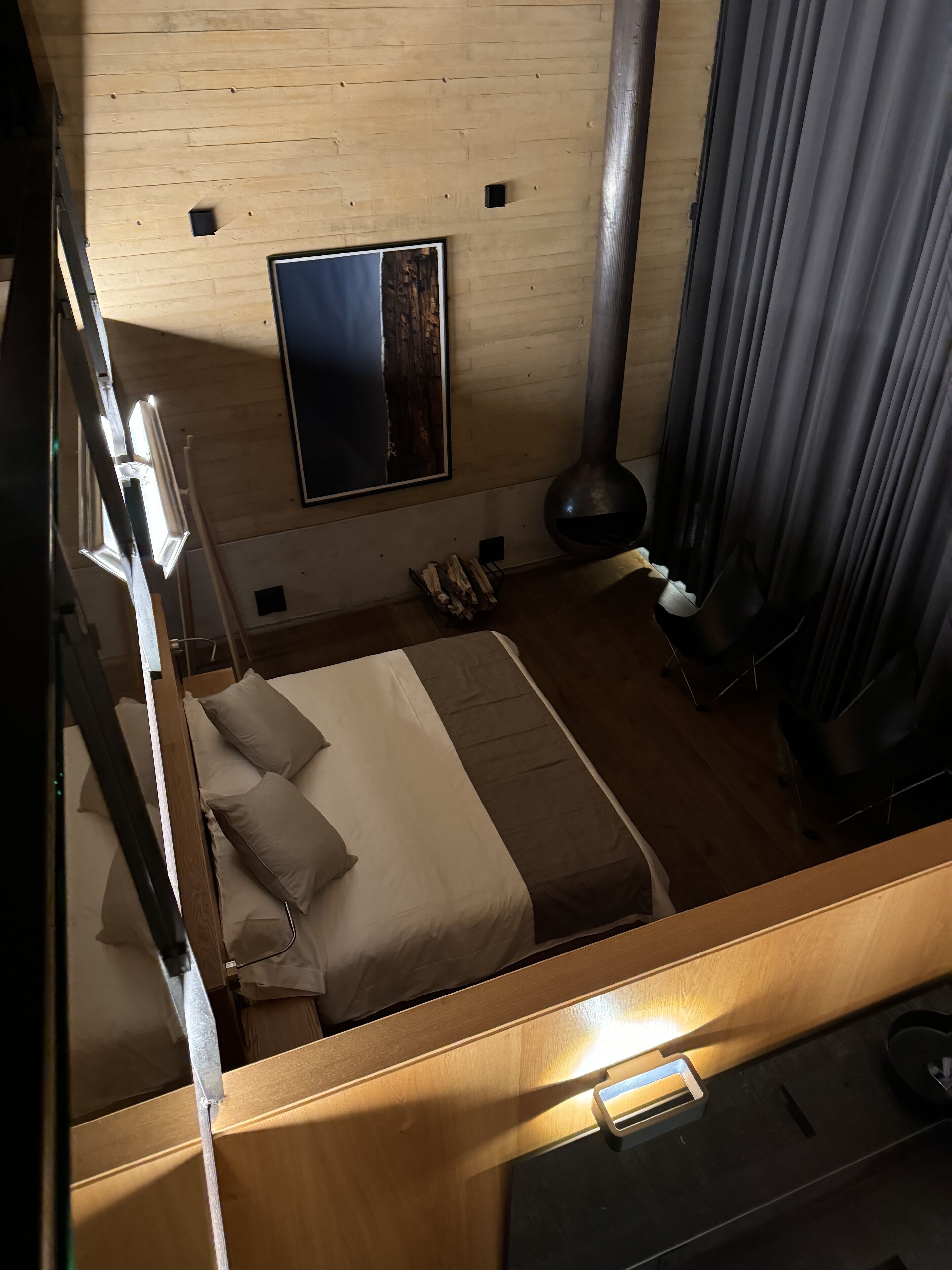 Una habitación de hotel moderna con suelo de madera, una cama con ropa de cama beige y marrón, un espejo grande en la pared, una lámpara negra y una ventana con cortinas.