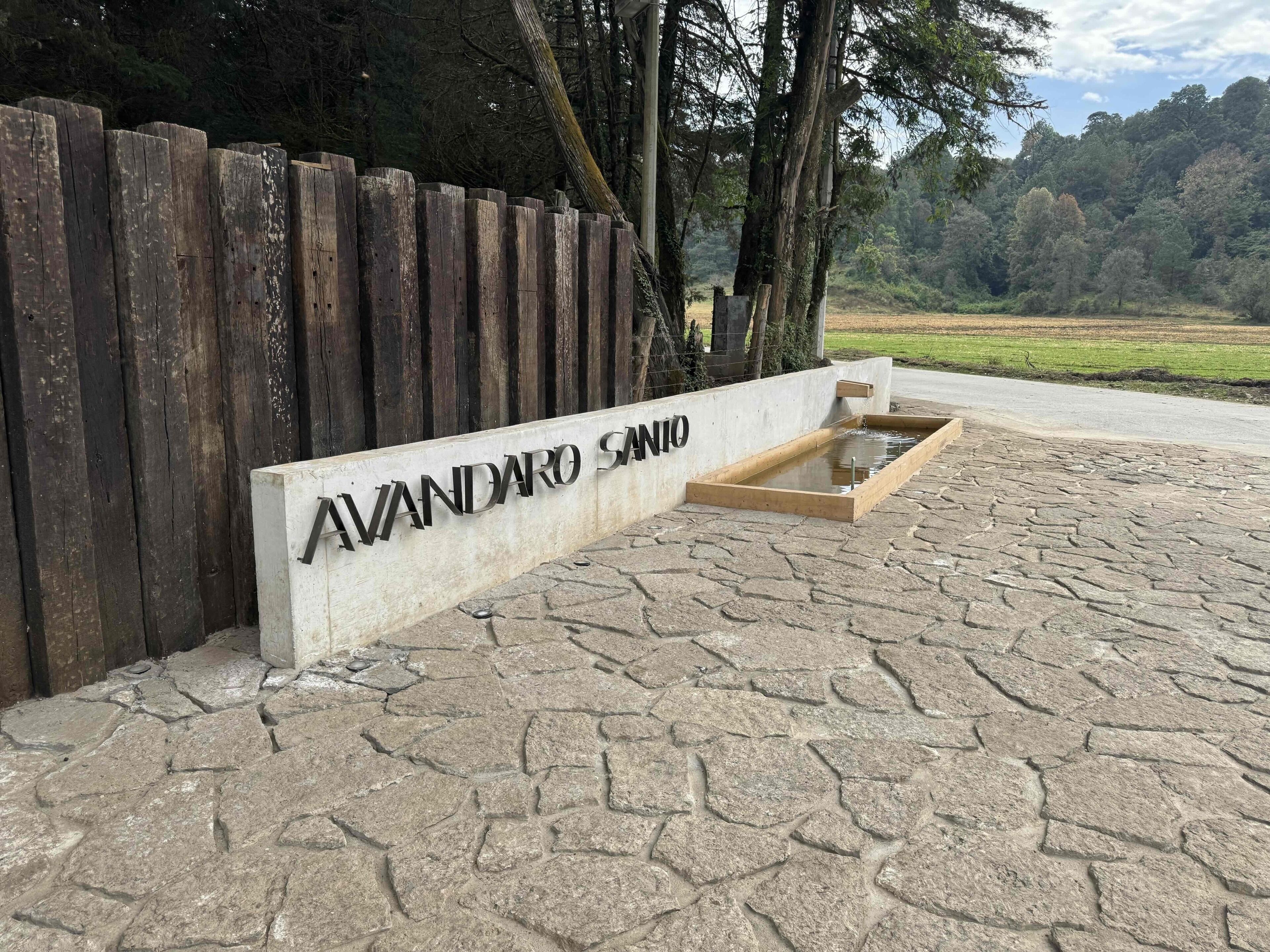 Une pancarte portant l’inscription « AVANDARO SANIO » est fixée à un mur en béton. On aperçoit en arrière-plan une clôture en bois et, au premier plan, une zone pavée de pierres.