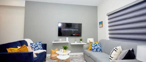 A modern living room with a grey sofa, a blue sofa, a small white table, and a TV mounted on the wall.
