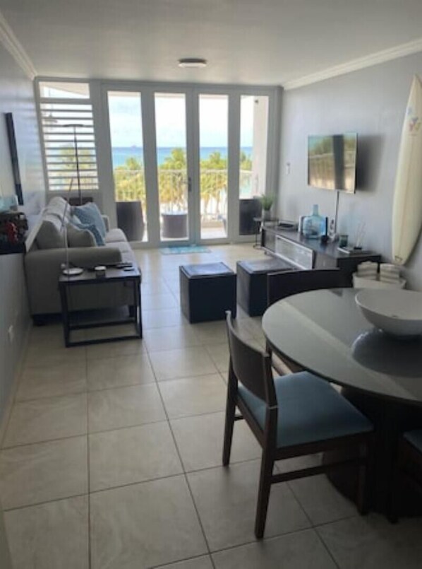 A modern living room with a sofa, coffee table, and dining area. There is a large sliding glass door leading to a balcony with a view of the ocean.