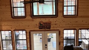 Une pièce spacieuse avec un plafond haut, des murs en bois et de grandes fenêtres. On y trouve un ventilateur de plafond, une enseigne indiquant « Bienvenue à Adventure Grace on the Rivers, fondée en 2023 », et une porte donnant sur l’extérieur.