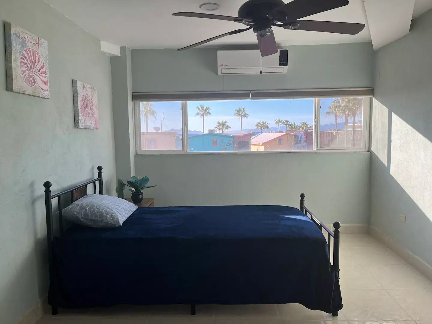 A bedroom with a bed, a ceiling fan, an air conditioner, and a window with a view of palm trees and buildings.