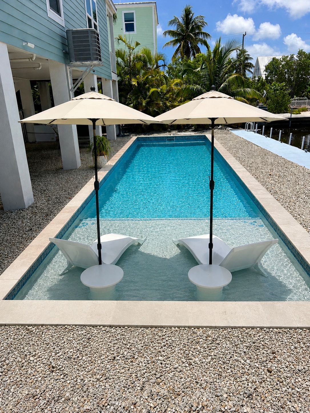 Piscina rectangular con dos sillas de descanso blancas y sombrillas, rodeada de guijarros, con un edificio y palmeras al fondo.