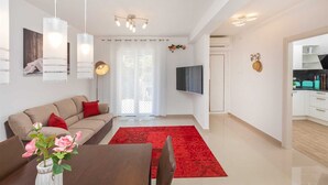 A modern living room with a sofa, a dining table, and a red rug. There is a TV mounted on the wall and a kitchen visible through an open door.