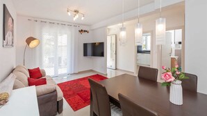 A modern living room with a sofa, red rug, dining table, and chairs. A TV is mounted on the wall, and there is a kitchen visible through an open door.