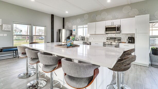 A modern kitchen with a large island, bar stools, stainless steel appliances, and decorative wall art. - Modern Farmhouse in Del Valle: Rural Setting! (Del Valle)