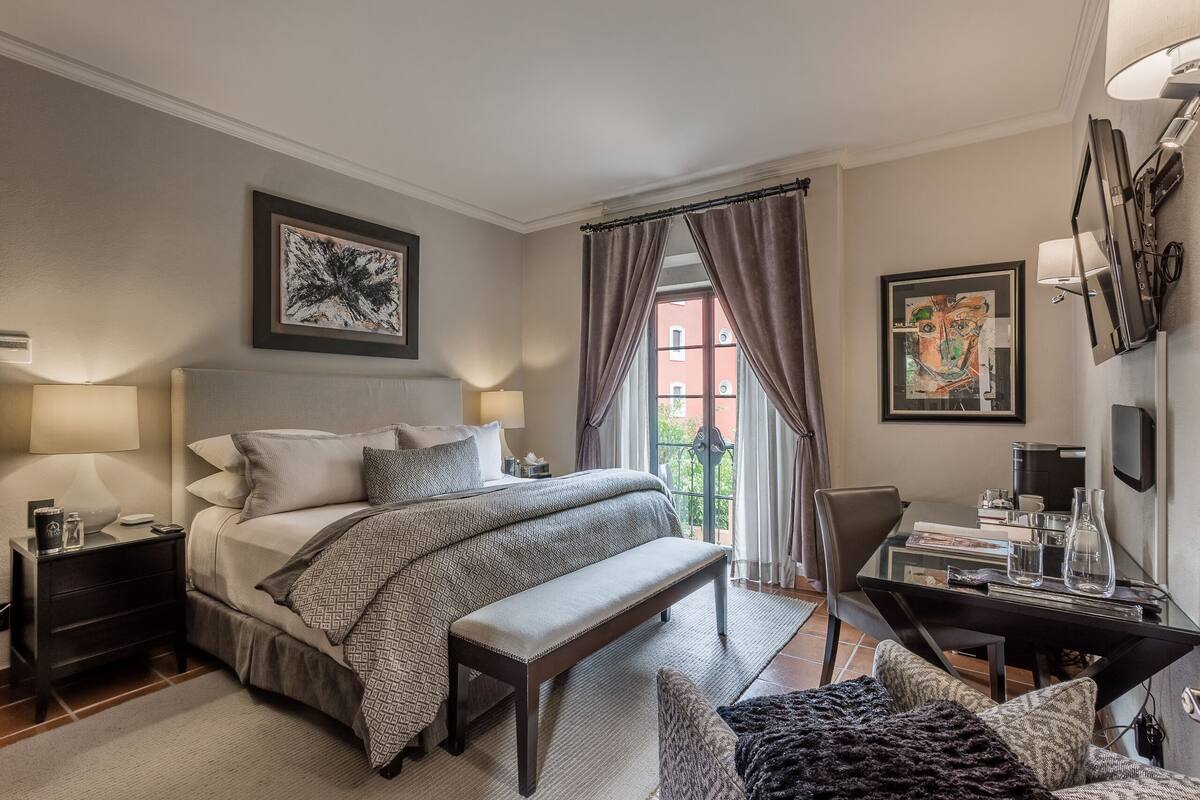 A bedroom with a large bed, bedside tables, a bench, a desk with a chair, and artwork on the walls.