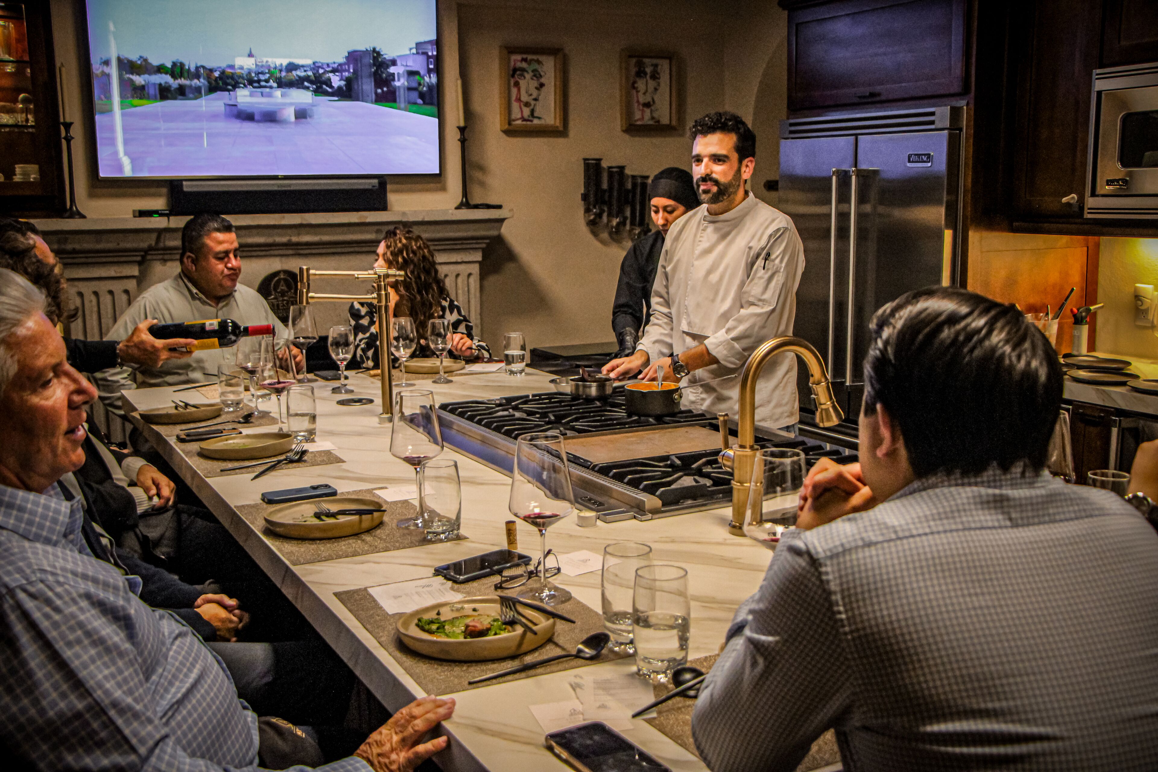 A group of people seated around a long dining table in a restaurant with a chef standing at the center.