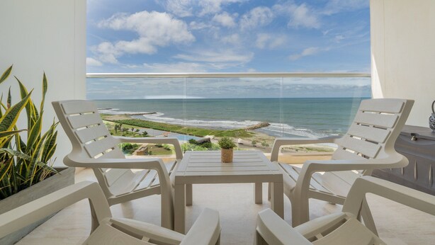 A balcony with two white chairs and a small table, overlooking a beach and ocean view.