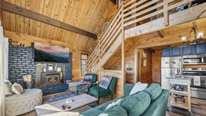 A cozy cabin interior with a stone fireplace, green upholstered seating, a kitchen with stainless steel appliances, and a wooden staircase. - Epic Views| 3 Pools!| Dog Friendly| Gatlinburg, GSMNP 10 Minutes (Gatlinburg)