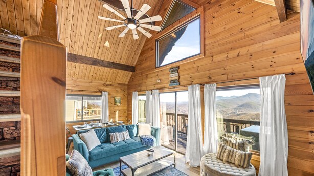 A cozy cabin interior with a wooden ceiling, a blue sofa, a round ottoman, and a view of the mountains through large windows. - Epic Views| 3 Pools!| Dog Friendly| Gatlinburg, GSMNP 10 Minutes (Gatlinburg)