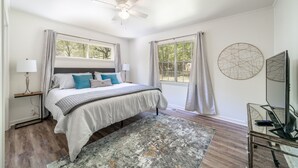 Une chambre à coucher comprenant un lit, des lampes de chevet, un ventilateur de plafond, une grande fenêtre avec des rideaux, un tableau rond décoratif et une table en verre avec une télécommande.