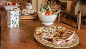 Une table en bois sur laquelle se trouve une assiette de pain grillé garnie d’herbes, un bol de légumes variés et une bouteille d’huile d’olive.