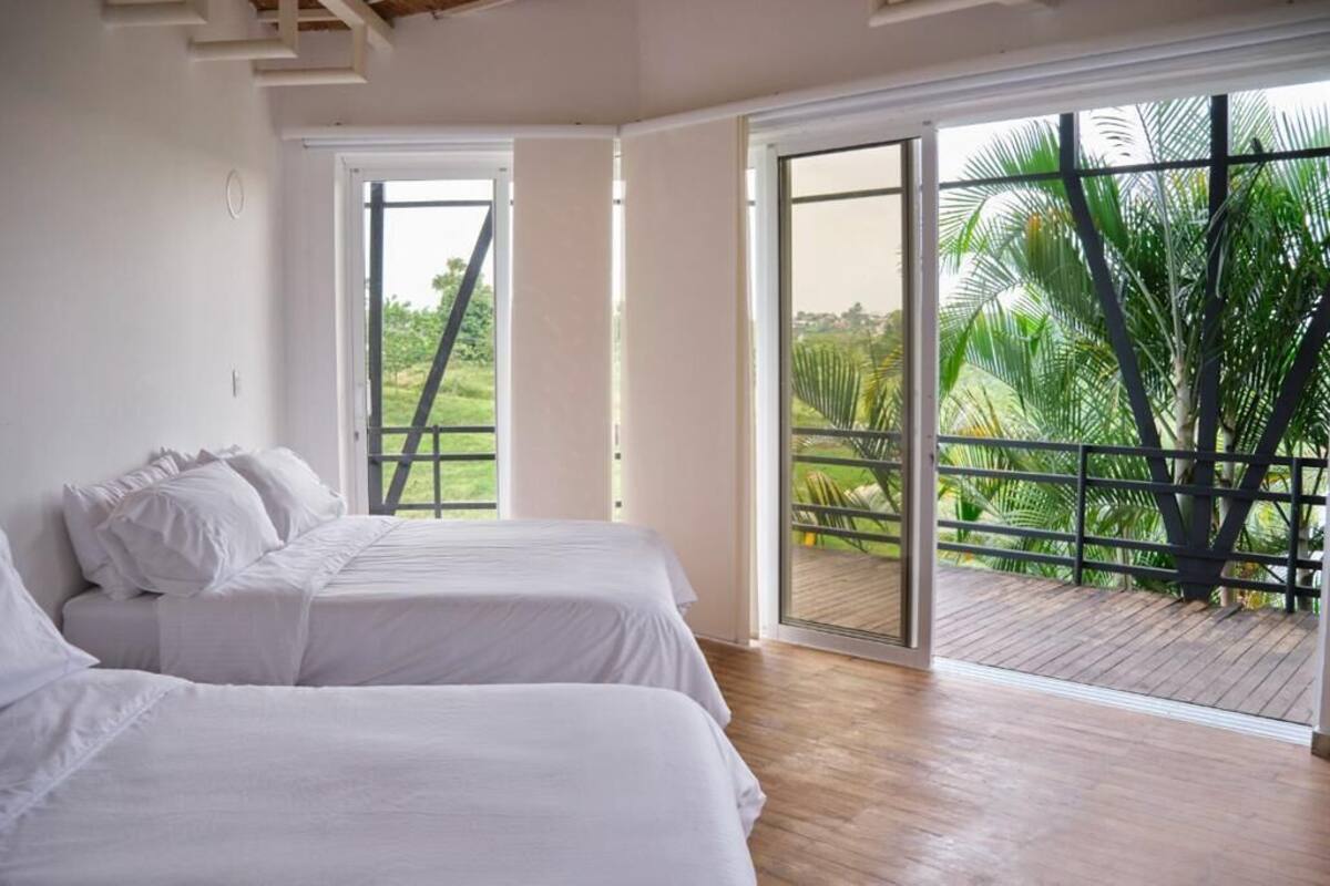 A room with two beds, large windows, and a view of greenery outside.