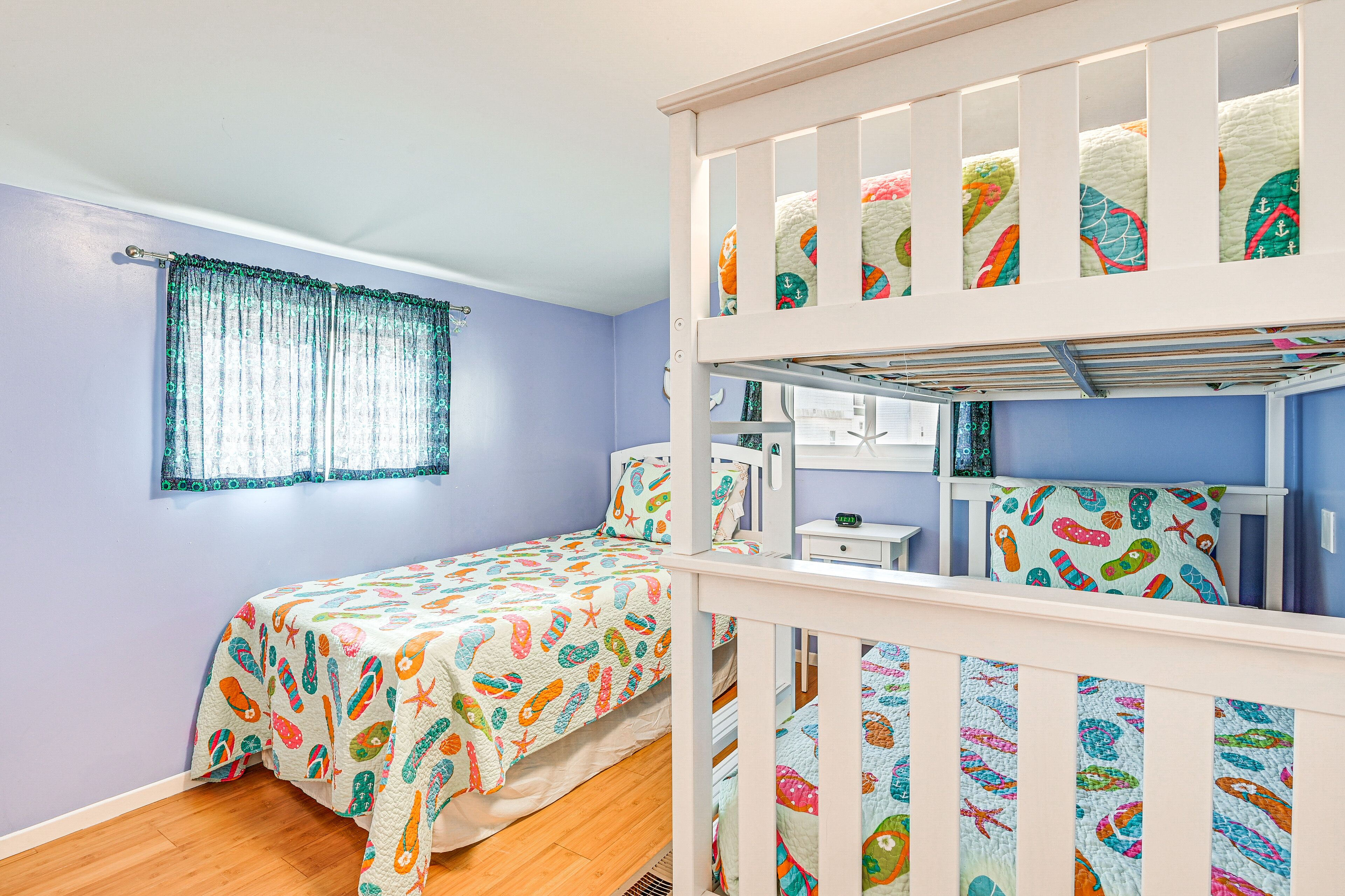 Habitación infantil con literas, cama con colcha de motivos de peces de colores, ventana con cortinas translúcidas y una mesita de noche con lámpara.