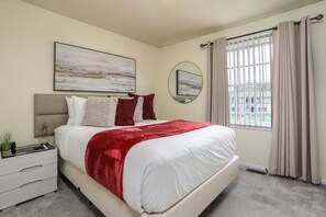 A bedroom with a bed, a nightstand, a mirror, and a window with curtains. - FamilyNest/King Bd/GameRoom (Royal Oak)