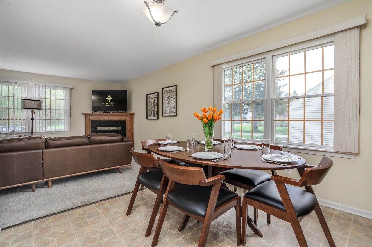Un coin repas avec une table en bois dressée pour six personnes, entourée de six chaises. On y trouve également un canapé en cuir marron et une cheminée sur laquelle est posée une télévision.