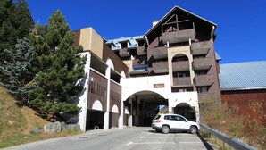 A multi-story building with a ski lift entrance, parking area, and a white car parked in front. - Studio 4/6pers, Terrasse, Jardinet, vue Imprenable sur la Muselle.50m des Pistes (Les Deux Alpes)