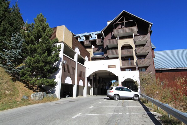 A multi-story building with a ski lift entrance, parking area, and a white car parked in front. - Studio 4/6pers, Terrasse, Jardinet, vue Imprenable sur la Muselle.50m des Pistes (Les Deux Alpes)