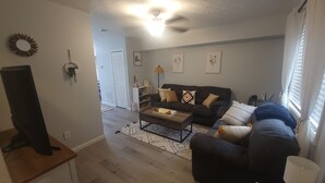Ein Wohnzimmer mit einem dunklen Sofa, einem Couchtisch und einem Fernseher auf einem Schrank. Eine Tür führt in einen anderen Raum, und ein Fenster ist mit Vorhängen drapiert.