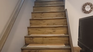 Eine Holztreppe mit ebener Oberseite und einer Handläufe auf der linken Seite, die zu einer Zwischenebene führt. Ein Flachbildfernseher ist an der Wand rechts neben der Treppe angebracht.