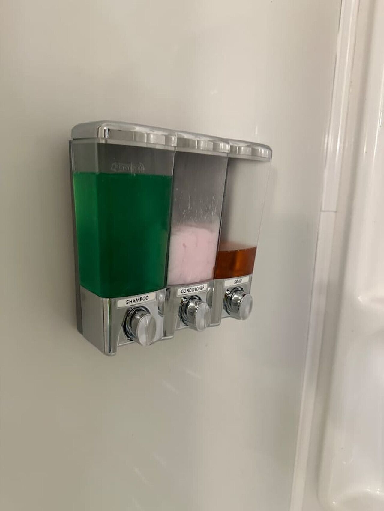 A wall-mounted dispenser with three compartments containing shampoo, conditioner, and soap.