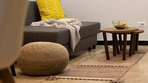 Un salon moderne avec un canapé, une table ronde en bois sur laquelle est posé un saladier de fruits, un pouf en fibres tressées et un tapis à motifs.