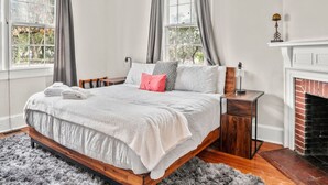 A bedroom with a large bed, a fireplace, and a view of trees through the windows. - Simplicity on Sims | 3BR 1BA | Downtown (Columbia)