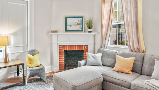 A living room with a grey sectional sofa, a small side table, a chair, a fireplace, and a window with curtains. - Simplicity on Sims | 3BR 1BA | Downtown (Columbia)