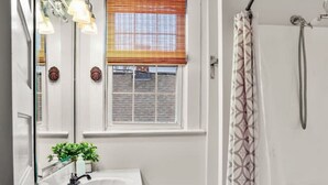 A bathroom with a white vanity, a window with blinds, a shower with a patterned curtain, and a small plant on the vanity. - Simplicity on Sims | 3BR 1BA | Downtown (Columbia)