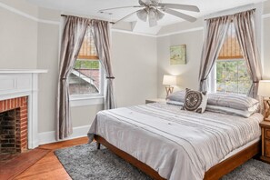 A bedroom with a bed, a fireplace, a ceiling fan, and a window with curtains.