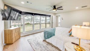 A bedroom with a large window, a ceiling fan, a TV mounted on the wall, a bedside table with a lamp, and a bed with a white comforter. - Grand Lakefront Home in Hot Springs w/ Boat Dock! (Hot Springs)