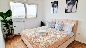 A bedroom with a bed, a window, a radiator, a plant, and two wall art pieces. - Charming Apartment With Panoramic Seine River & Eiffel Tower Views (Ivry-sur-Seine)