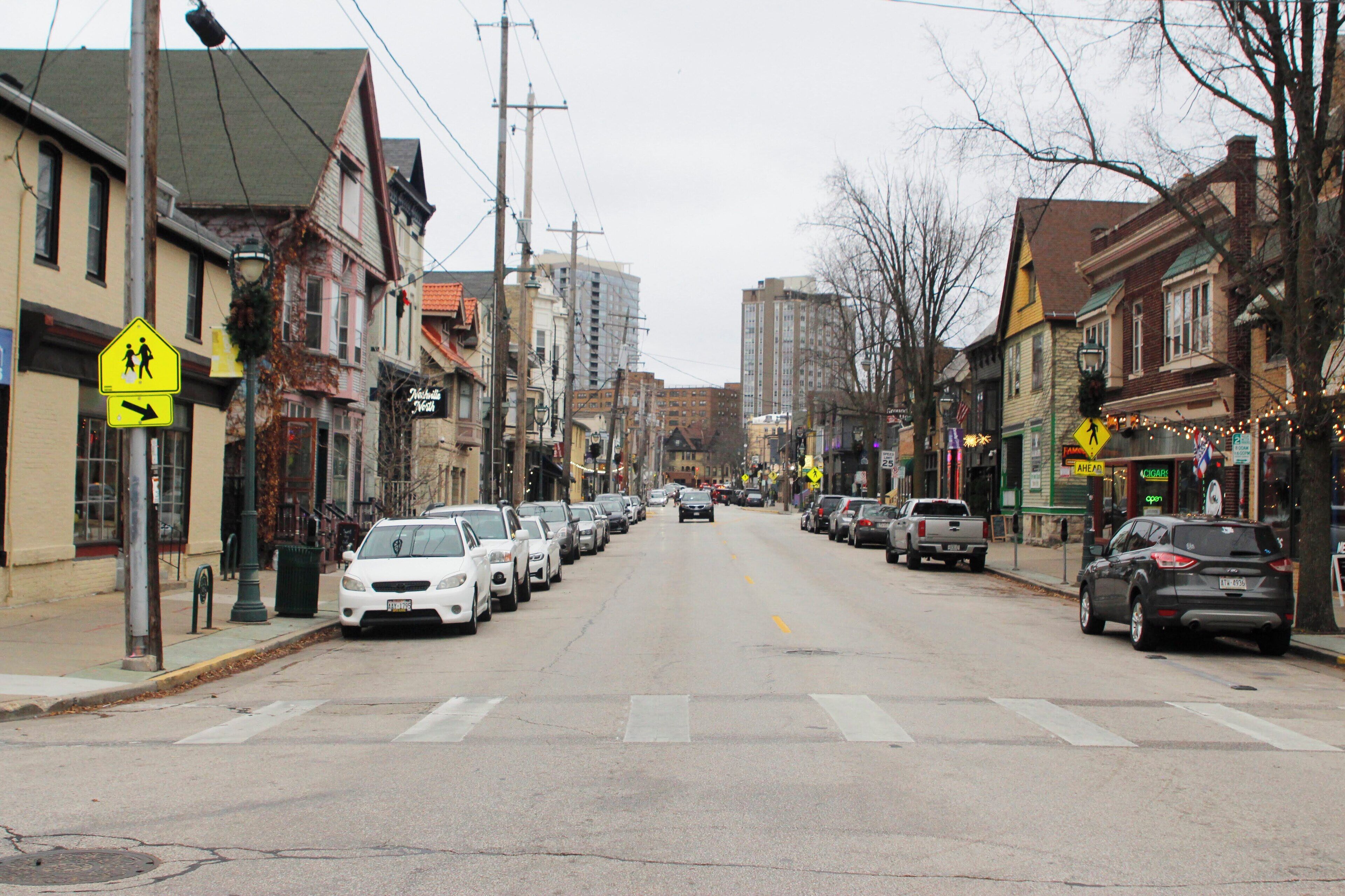 A street lined with parked cars and buildings, including a pedestrian crossing sign.