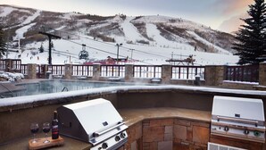 Un espace extérieur aménagé pour les barbecues, avec une cheminée en pierre, un jacuzzi et une vue sur un télésiège et des montagnes enneigées.