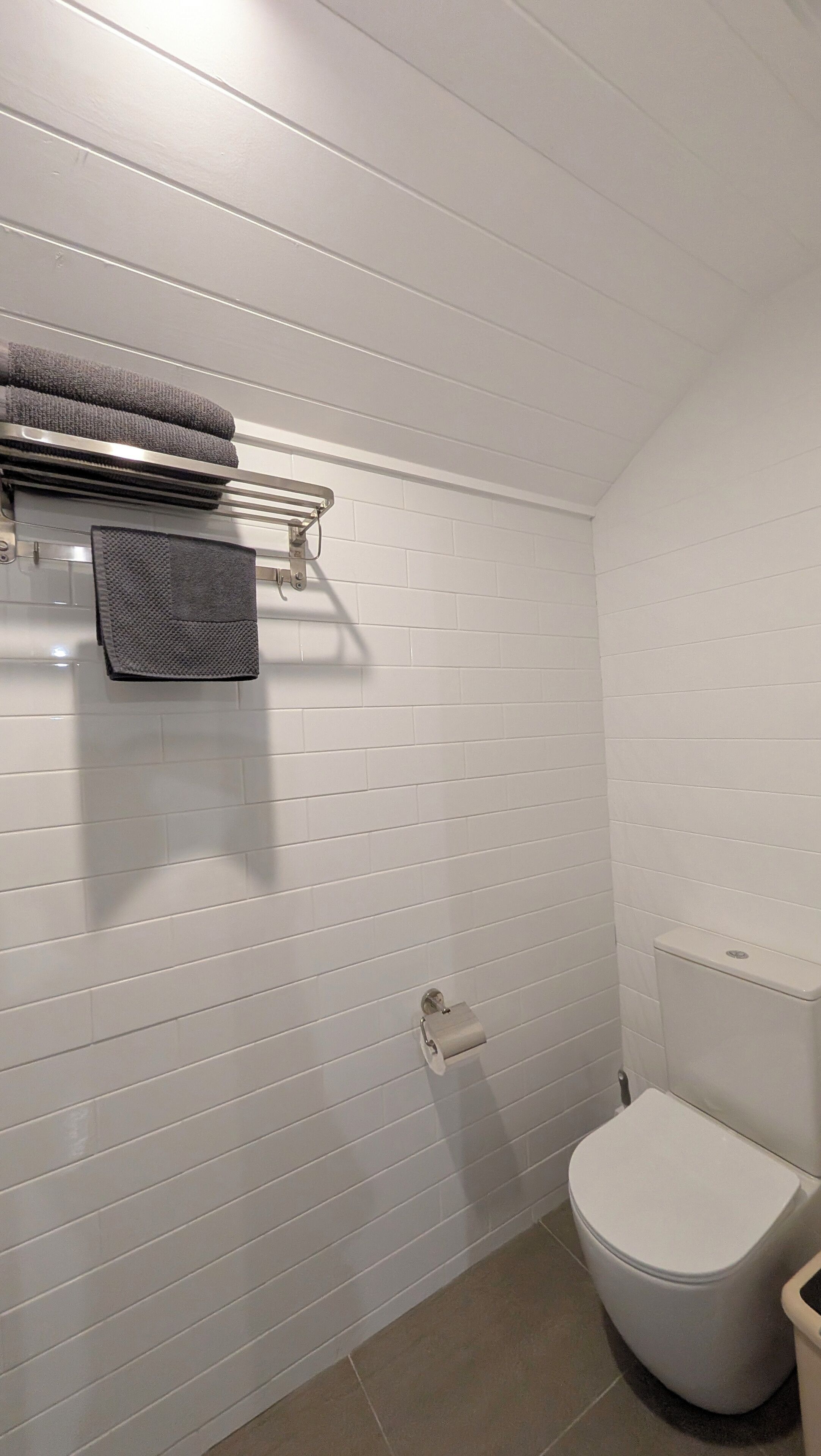 A bathroom with white tiled walls, a white toilet, a towel rack with folded towels, and a toilet paper holder.
