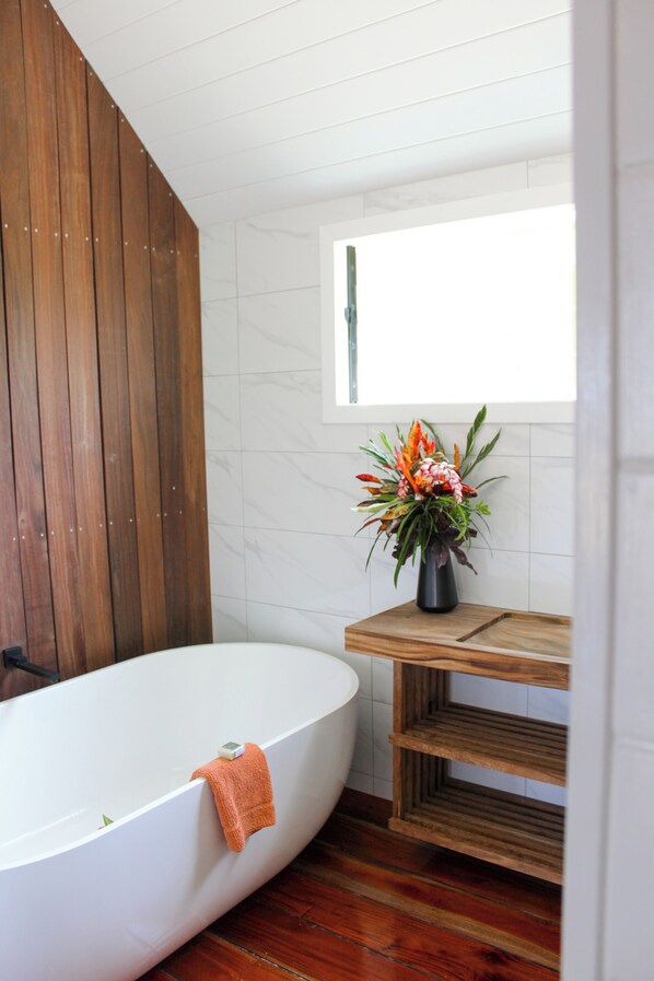 A bathroom with a wooden slatted wall, a white bathtub, a wooden vanity, and a window with a view. - Bombua Beach House (Luganville)