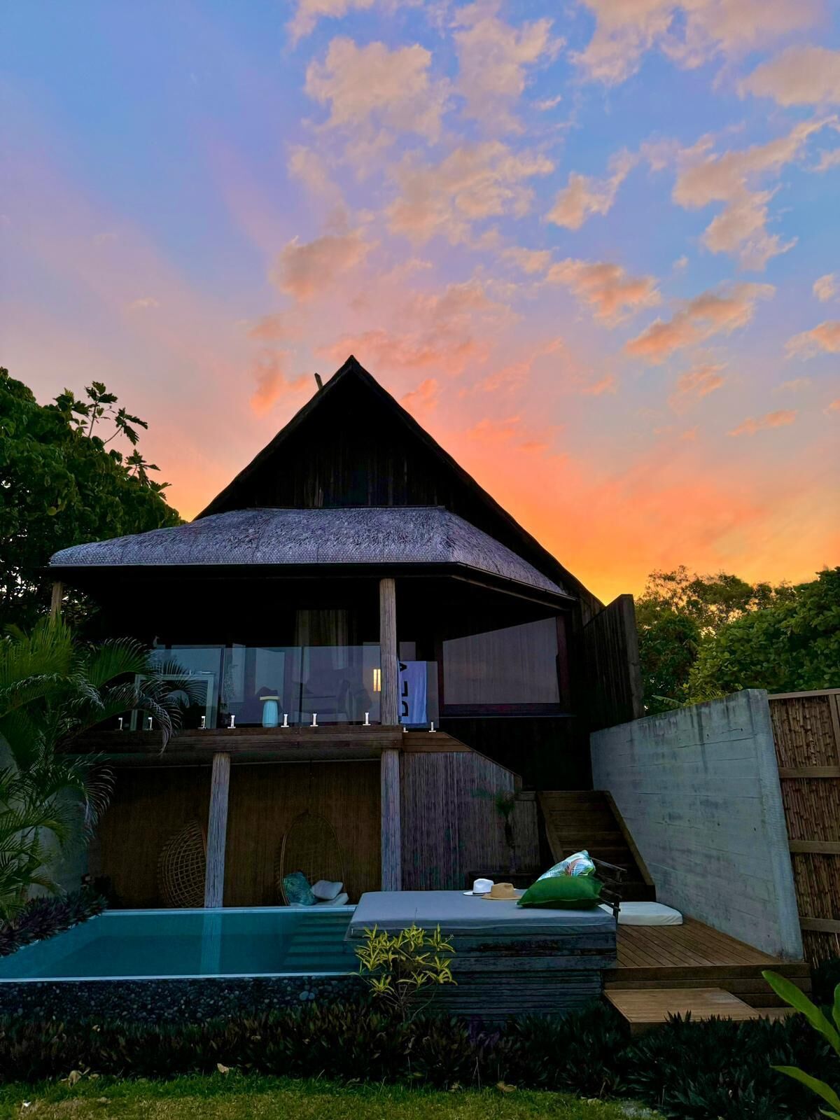 Una casa moderna con un amplio balcón de vidrio, piscina y abundante vegetación.