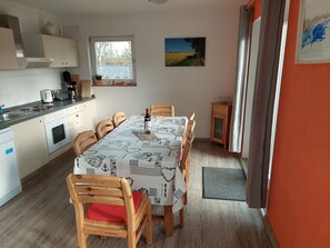 A compact kitchen with a dining area, featuring a table set for two, wooden chairs, a small cabinet, and a window with a view of a field. - Erholungszeit im Haus Ostseeblick (Kalkhorst)