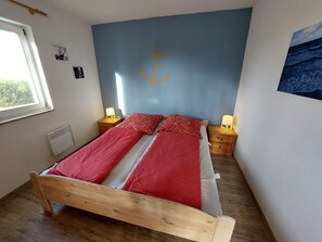 A bedroom with a wooden bed, red bedding, two bedside tables with lamps, a window, and a blue accent wall with an anchor design. - Erholungszeit im Haus Ostseeblick (Kalkhorst)