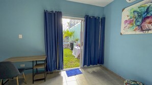 A room with a sliding glass door leading to a garden, a small table, a chair, and a colorful painting on the wall.