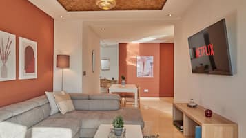 A modern living room with a sectional sofa, a coffee table with a plant, a TV mounted on the wall, and a dining area in the background.